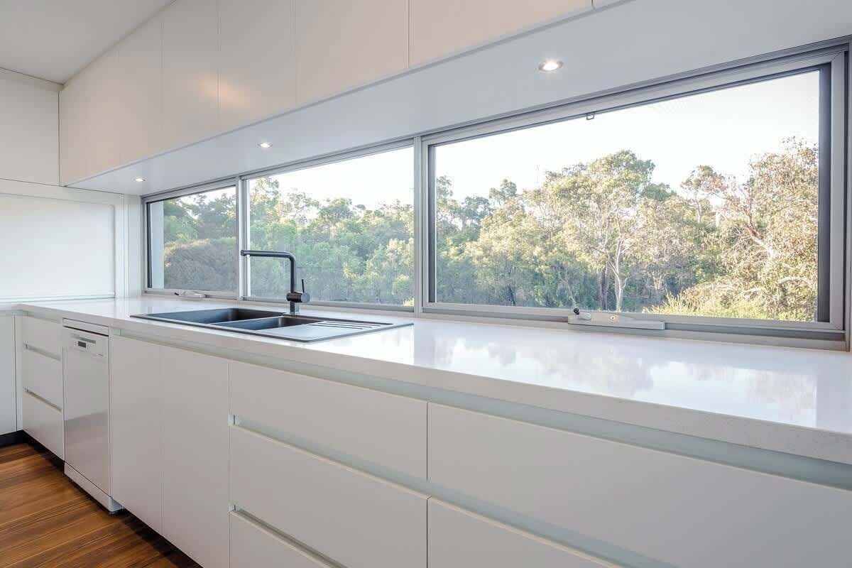 Una cocina moderna con un ventanal grande, muebles blancos y un lavamanos de acero inoxidable.