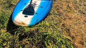 Un kayak bleu, avec une pagaie et un gilet de sauvetage rouge, est posé sur une rive herbeuse au bord d’un lac.