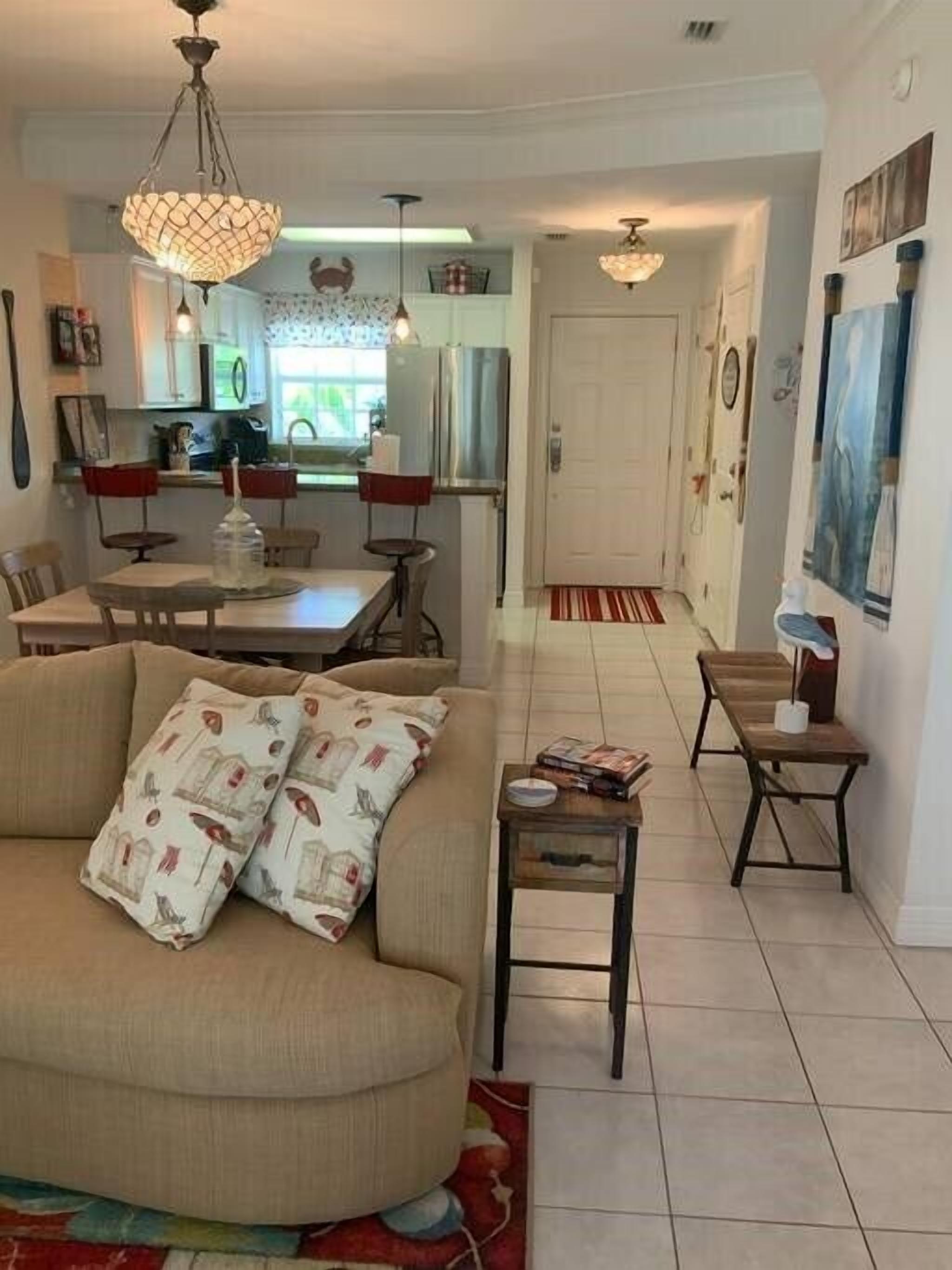 A living room with a sofa, a coffee table, and a dining area with a table and chairs. There is a kitchen in the background with cabinets and a refrigerator.