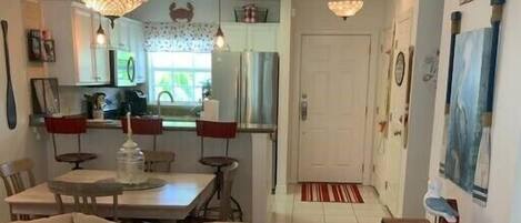 A living room with a sofa, a coffee table, and a dining area with a table and chairs. There is a kitchen in the background with cabinets and a refrigerator.