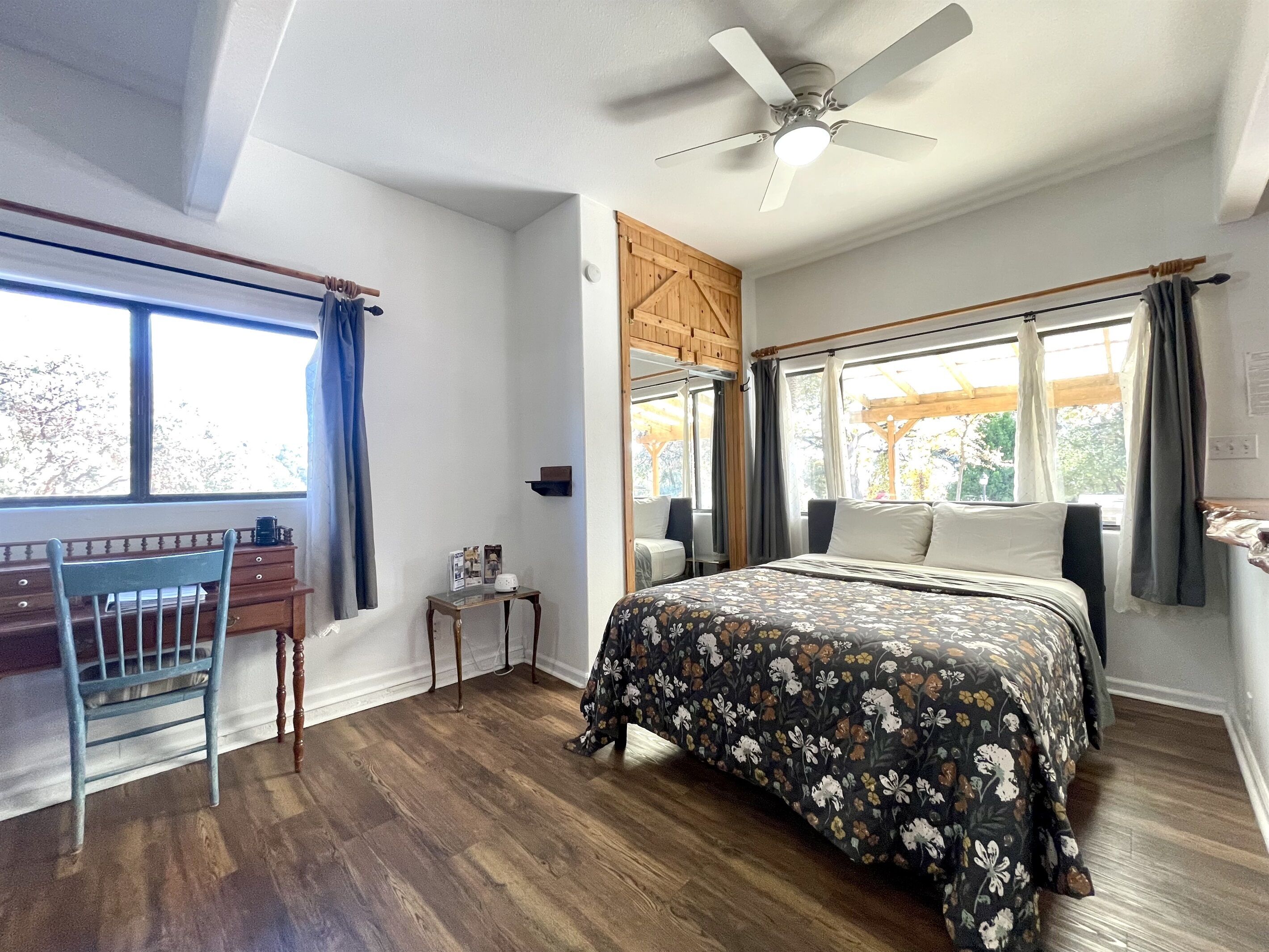 Ein Schlafzimmer mit Bett, Schreibtisch, Stuhl, Deckenventilator und großen Fenstern.