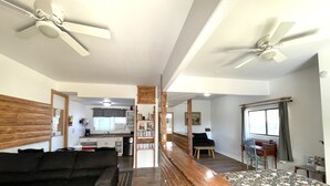 Een kamer met een bed, een slaapbank, een houten hoofdbord, een plafondventilator, een raam en een kitchenette op de achtergrond.
