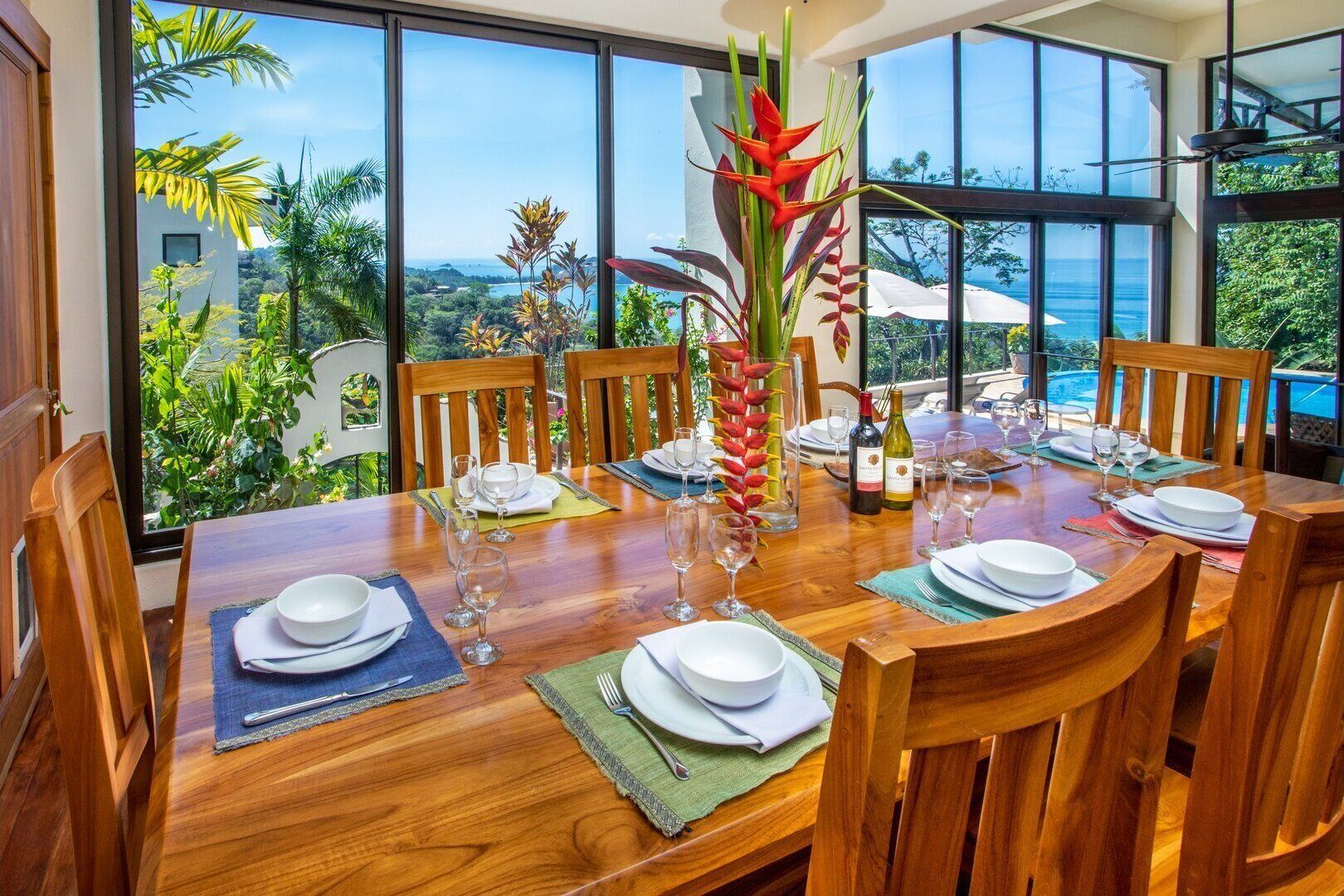 A dining table set for six with a view of the ocean and lush greenery.