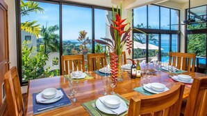 Mesa para seis personas dispuesta con vista al océano y abundante vegetación.