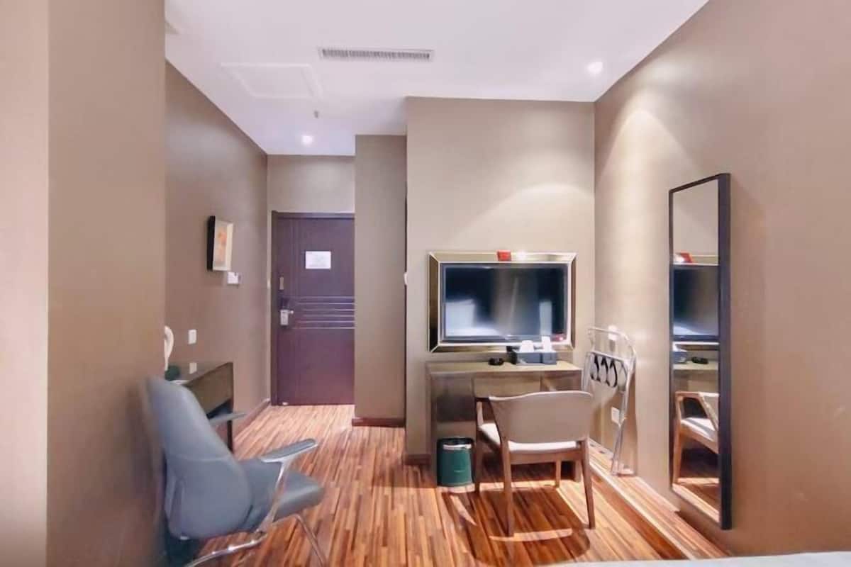 A hotel room with a wooden floor, a desk with a TV, a chair, a mirror, and a door.