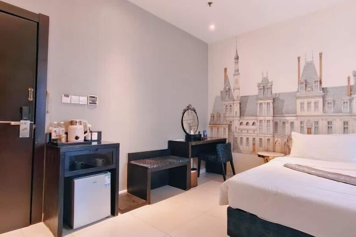 A hotel room with a bed, a desk, a chair, a small refrigerator, and a wall mural.