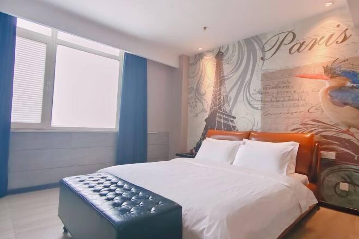 A hotel room with a bed, a window with blinds, a wall with a Paris-themed mural, and a clock.