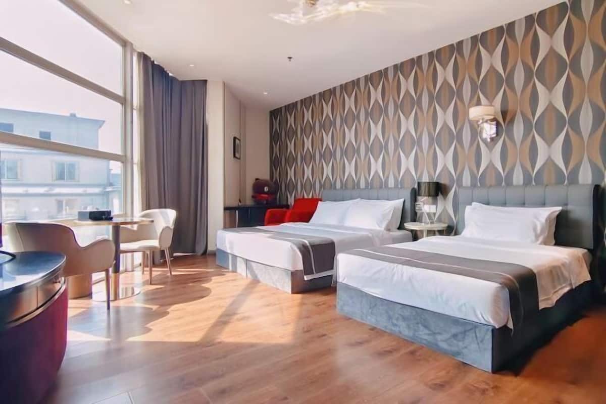 A hotel room with two beds, a desk, chairs, and a view of buildings.