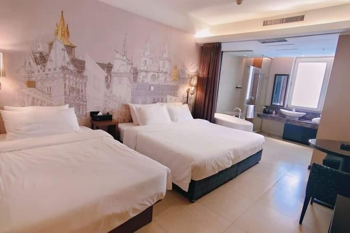 A hotel room with two beds, a bathroom with a sink and mirror, and a view of a building through the window.