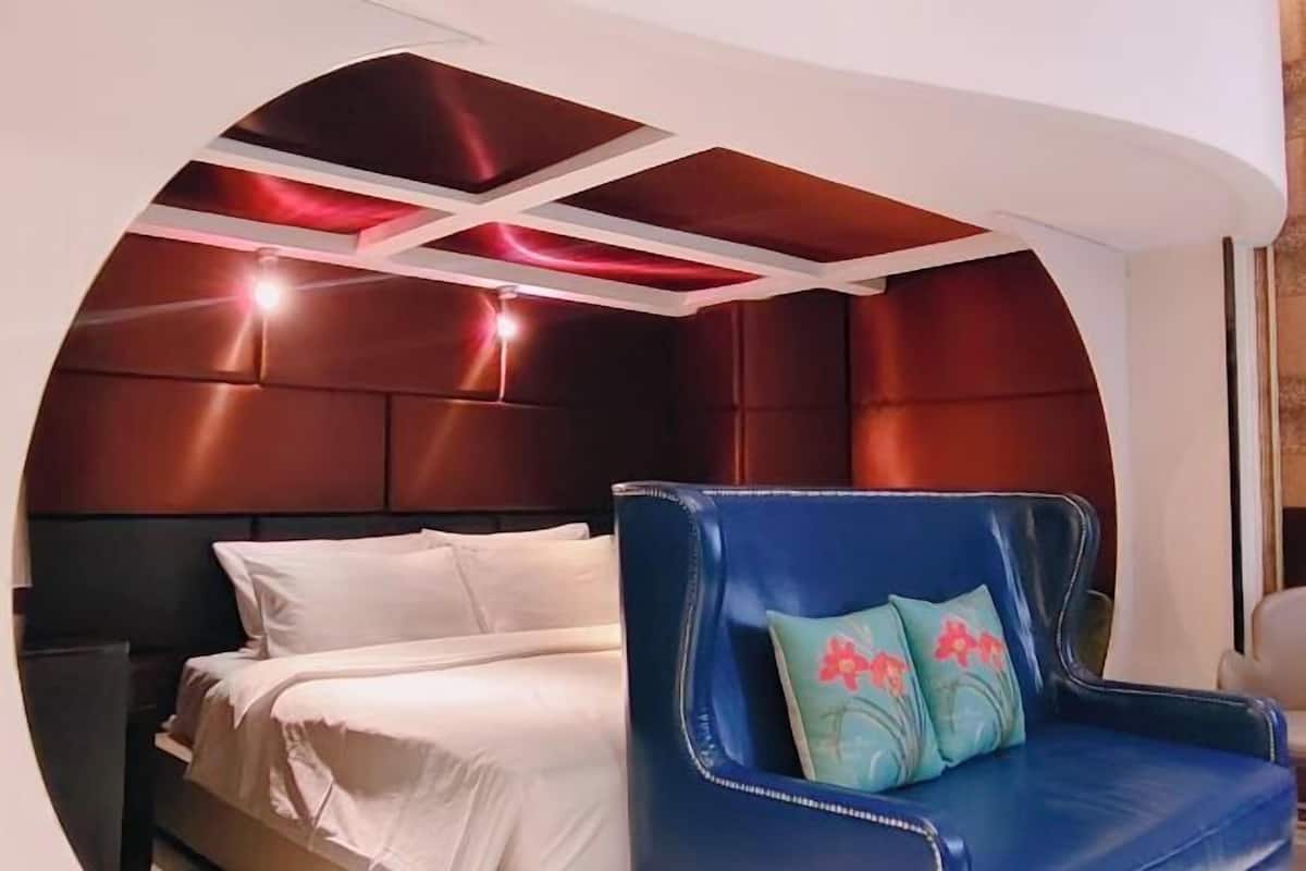 A room with a bed, a blue armchair with floral cushions, and a unique ceiling design.