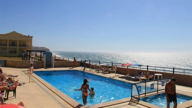 Uma área de piscina com vista para o oceano, cadeiras de descanso e um edifício ao fundo.