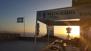 A seaside restaurant with outdoor seating, a sign indicating it is a cafe, and a view of the sunset over the ocean. - Praia Azul Hotel Apartamento (Torres Vedras)