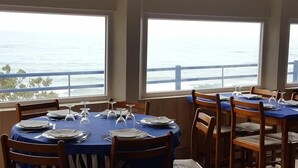 Restaurante com mesas redondas preparadas para refeições, grandes janelas com vista para o mar e cadeiras de madeira.