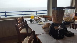 Mesa de café da manhã com vista para o mar, uma cafeteira e um copo de suco de laranja.
