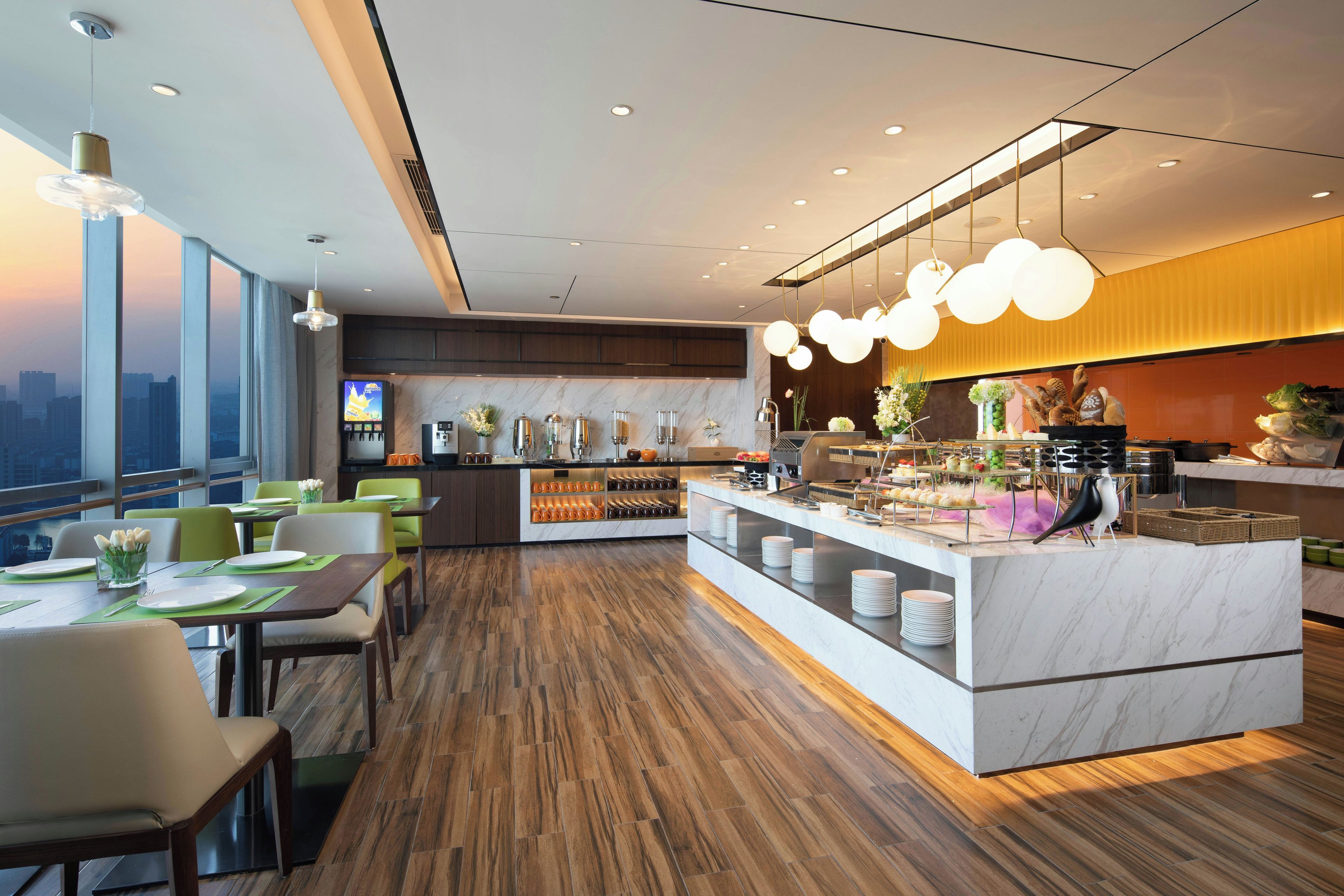 A modern hotel dining area with a central buffet, seating arrangements, and a city view.