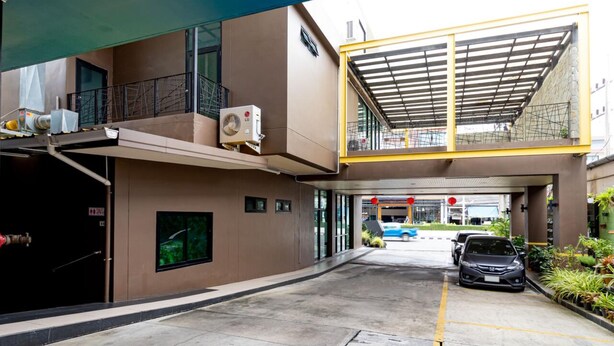 A modern building with a covered entrance, a visible air conditioning unit, and a parking area with cars. - Udee Living Place (Hat Yai)