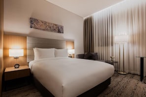 A hotel room with a large bed, two bedside tables with lamps, a nightstand, a chair, a small table with a vase, and a large window with curtains.