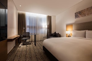 A hotel room with a large bed, a desk with a chair, a small table, and a lamp.