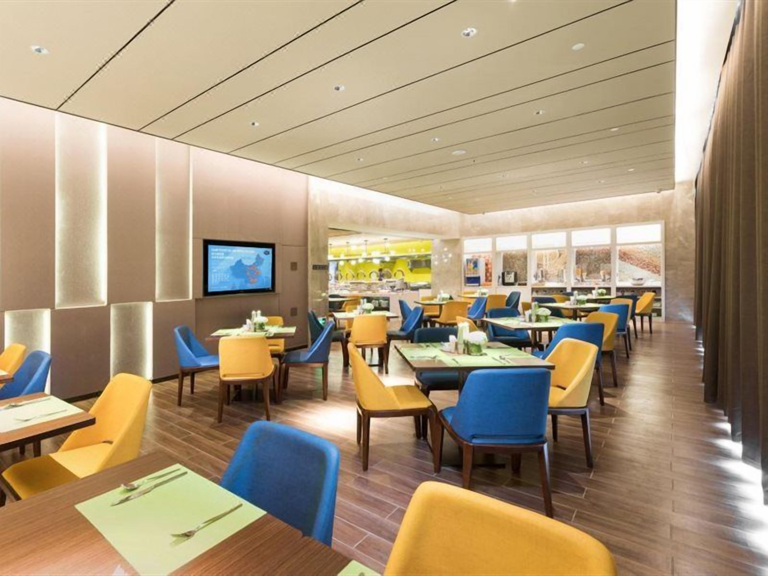 A modern dining area with multiple tables and chairs, a flat-screen TV mounted on the wall, and a view of the outside through large windows.