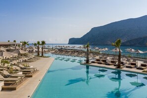 Zona de piscina en un resort con tumbonas y sombrillas, con vistas a una playa y montañas.