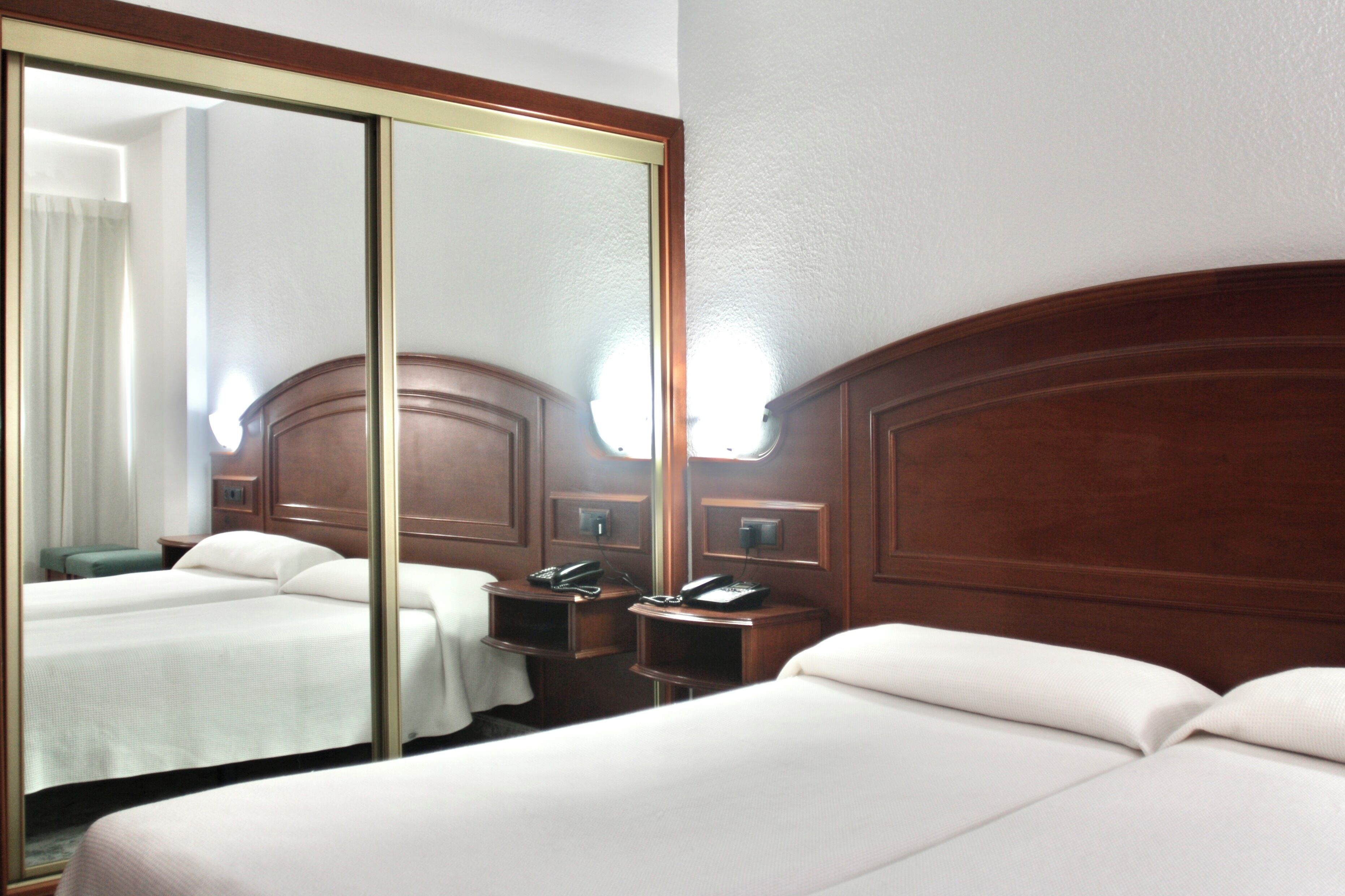 A hotel room with two beds, a large mirror, wooden headboard, bedside tables with lamps, and a telephone.