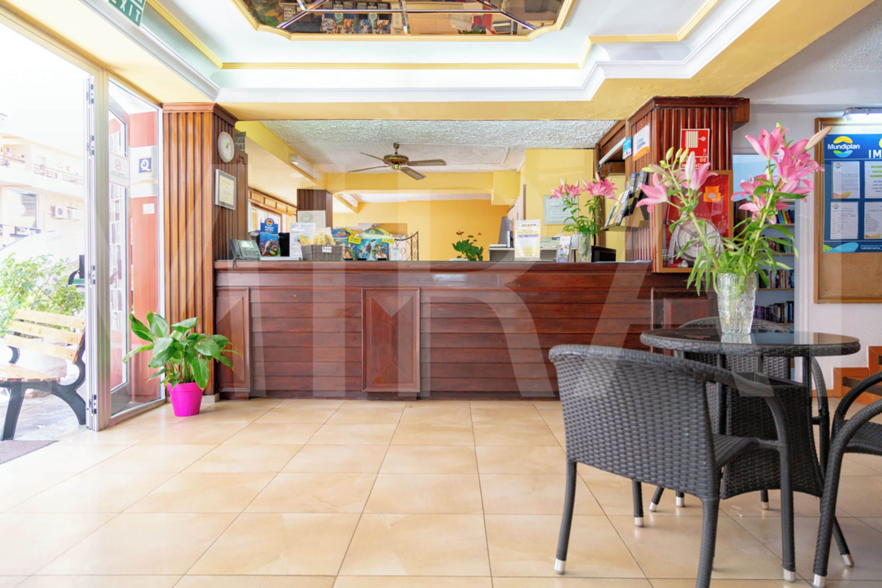 A hotel lobby with a reception desk, a ceiling fan, and a dining area with chairs and a table.
