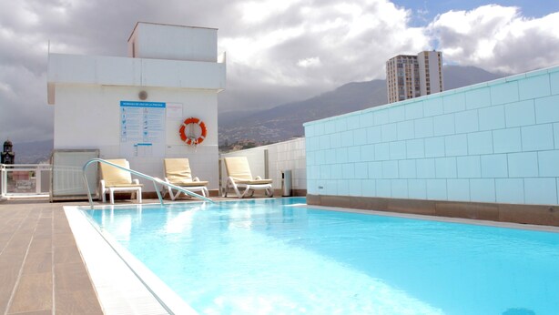 Una piscina en la azotea con sillones de descanso y un flotador salvavidas, rodeada por un edificio blanco y vistas a la montaña.