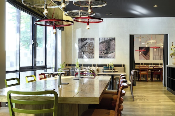 A modern dining area with a long wooden table, multiple chairs, and pendant lights. - Leonardo Hotel Nürnberg City Center (Nuremberg)