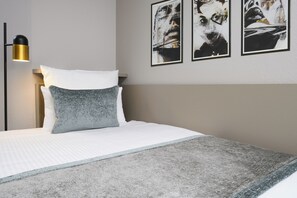 Una cama bien hecha con edredón blanco y almohada gris con textura, una lámpara de pie negra y dorada, y tres cuadros en la pared.