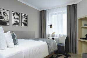 A hotel room with a bed, a chair, a small table with a vase, and a shelf with a cup. - Leonardo Hotel Nürnberg City Center (Nuremberg)