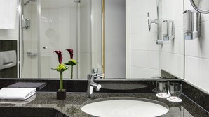 Un baño moderno con encimera de granito, un lavabo blanco, cabina de ducha de vidrio y un espejo con marco cromado.