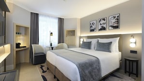 Une chambre d’hôtel avec un grand lit, des tables de chevet, un bureau, une chaise, une petite table avec une lampe et une fenêtre avec des rideaux.