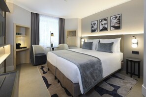 Habitación de hotel con una cama grande, mesitas de noche, un escritorio, una silla, una pequeña mesa con lámpara y una ventana con cortinas.