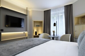 A hotel room with a bed, a TV, a desk with a chair, a small table with a vase, and a wardrobe.