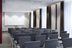 A conference room with rows of grey chairs, a projector screen, and a presentation table. - Leonardo Hotel Nürnberg City Center (Nuremberg)