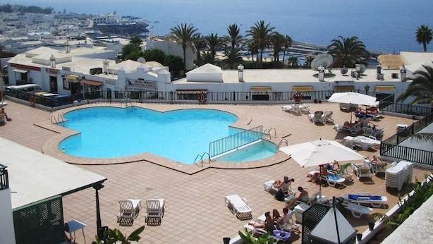 A rooftop pool with a curved design, surrounded by lounge chairs and umbrellas, overlooking a coastal town and the sea. - Los Pueblos Apartments (Tías)