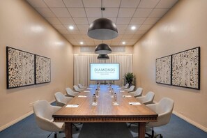 Un salón de conferencias con una larga mesa, sillas, una pantalla de proyector y detalles decorativos en la pared.