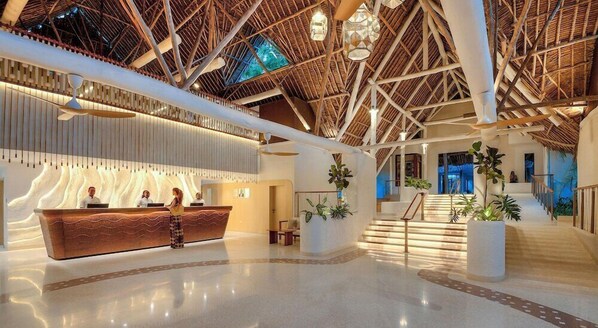 A reception area with a wooden reception desk, a high ceiling with a thatched roof, and a staircase leading to upper levels.