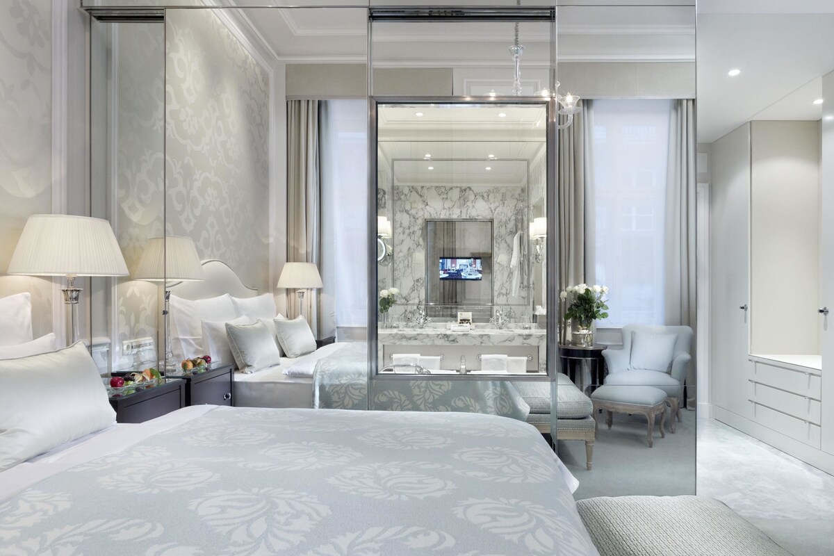 A modern hotel room with a large bed, a glass partition, a sitting area, a bathroom with a mirror, and a television.