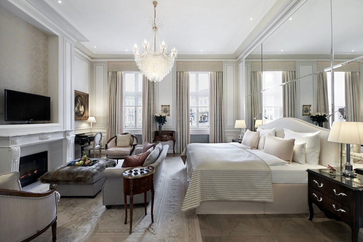 A spacious bedroom with a large bed, a sitting area with a sofa and armchairs, a fireplace, and a television.