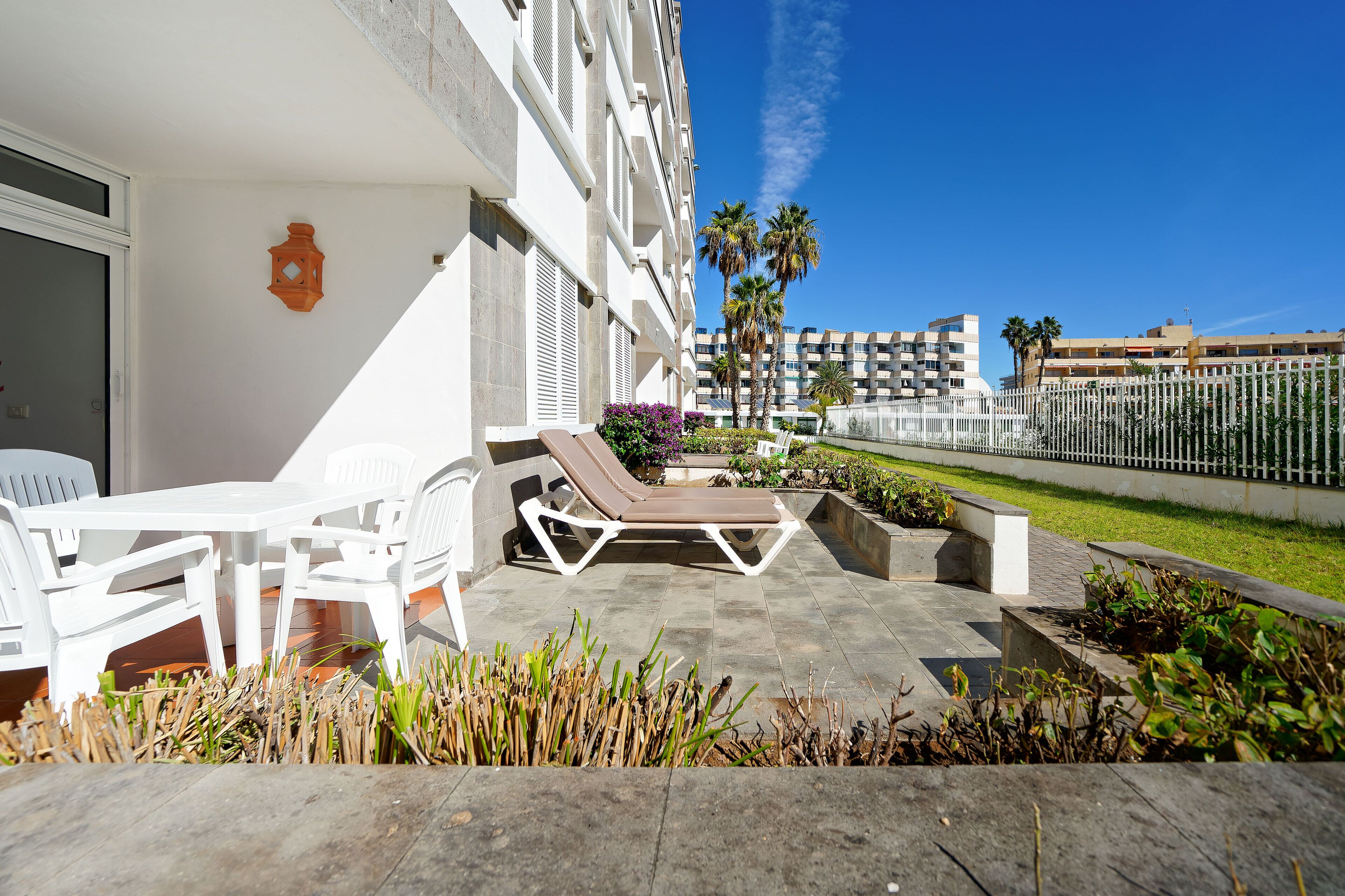 A hotel patio with white lounge chairs and a table, a small garden area with plants, and a clear view of the surrounding buildings and palm trees.