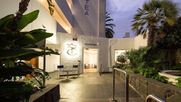 An entrance to a hotel with a reception desk, a sign with a symbol, and a stone pathway leading to the entrance. - TC Apartamentos Dorotea (San Bartolomé de Tirajana)