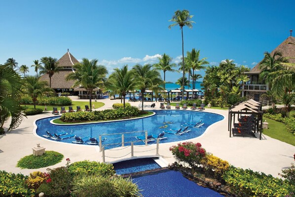 Um resort com uma piscina grande, espreguiçadeiras, palmeiras e um prédio com telhado de palha.