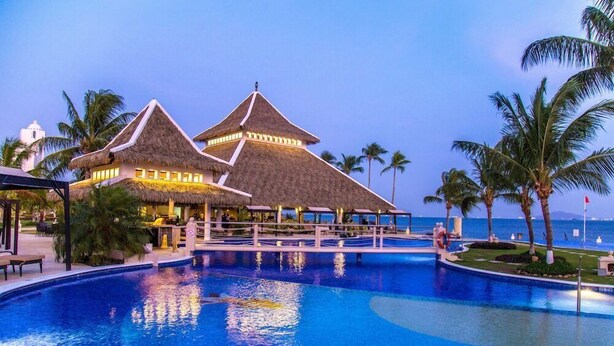 A resort pool area with thatched-roof structures, palm trees, and a clear view of the ocean. - Dreams Playa Bonita Panama (Veracruz)