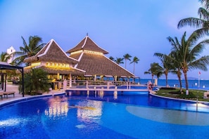 Ein Poolbereich in einem Resort mit strohgedeckten Gebäuden, Palmen und freiem Blick auf den Ozean.
