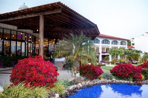 Um edifício com entrada coberta, piscina e jardins ajardinados com flores vermelhas.