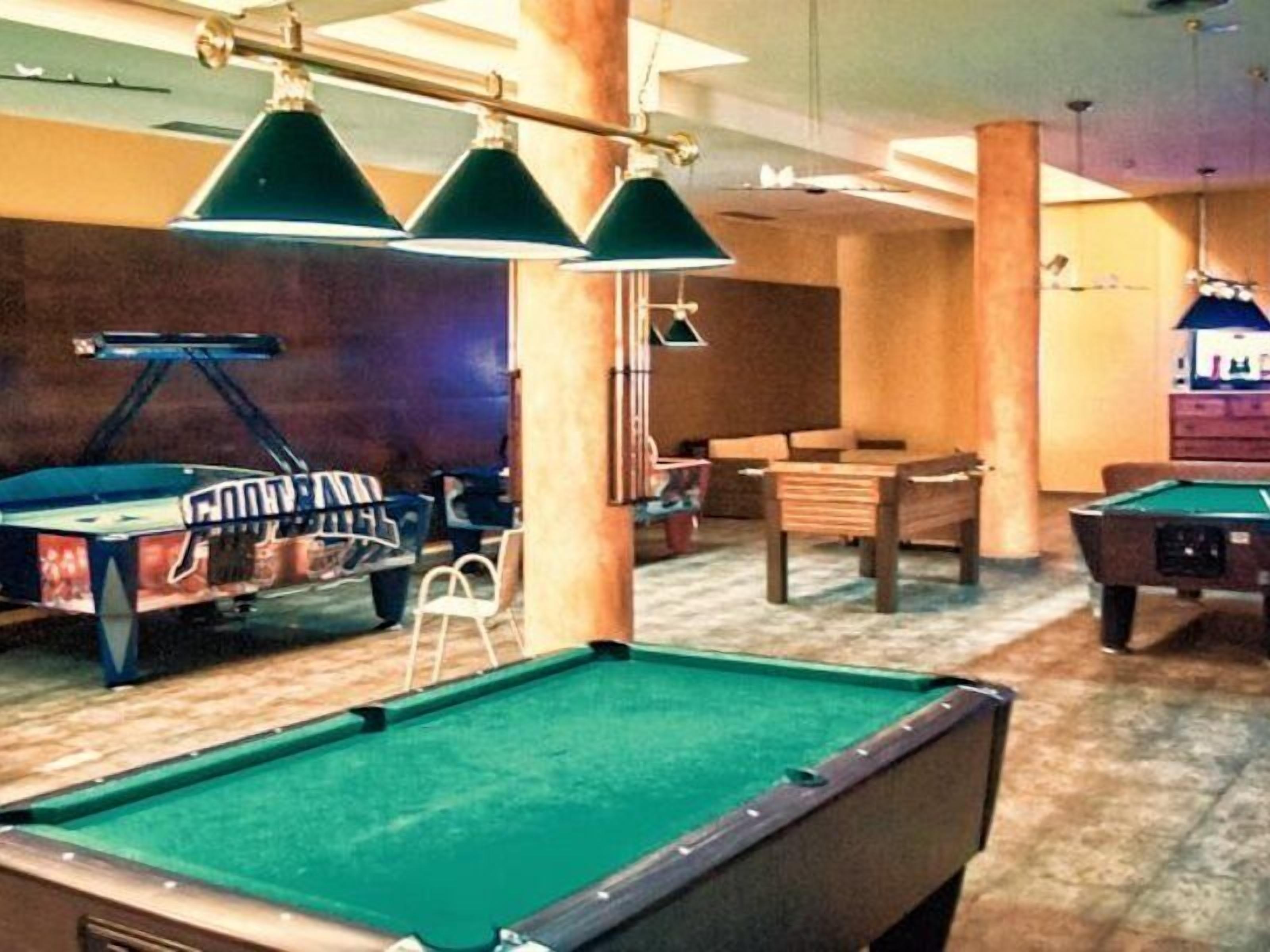 A pool table with a green felt surface, a pool cue, and a billiards rack with balls. There are two chairs and a small table in the background.