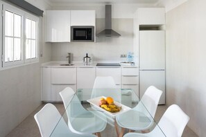 A modern kitchen with white cabinets, a microwave, a stainless steel range hood, and a refrigerator. There is a glass dining table with chairs and a fruit plate on it. - Hyde Park Lane Villas (Tías)