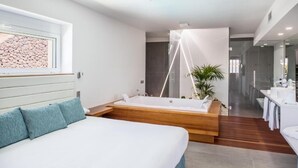 A modern bedroom with a large bed, a wooden platform bathtub, a small bedside table, and a large window with a view of rocks. - Hyde Park Lane Villas (Tías)