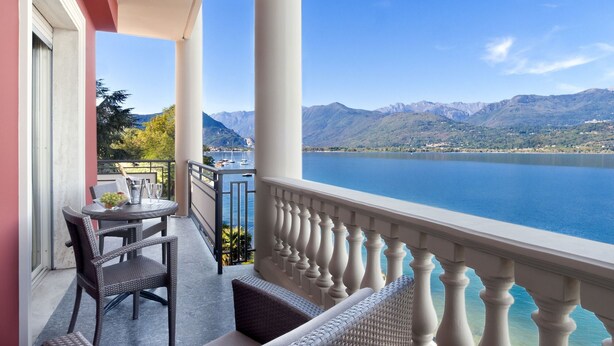 A balcony with wicker furniture, a table with a glass of wine, and a view of a lake and mountains. - Splendid (Baveno)